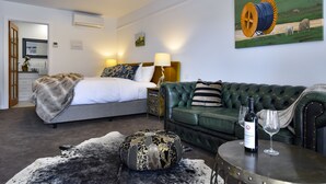 Una habitación de hotel con una cama, un sofá verde, una mesita con una botella y un vaso, y un cuadro en la pared.