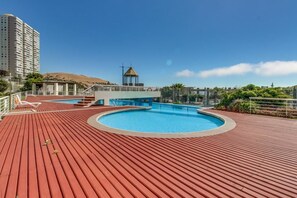 A rooftop swimming pool with a wooden deck, a lifeguard tower, and a view of a cityscape with a hill.
