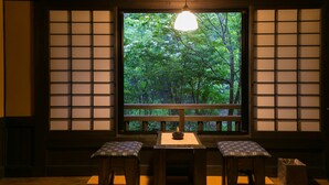 Ett traditionellt japanskt rum med tatamimattor på golvet, ett lågt bord med ett ljus och utsikt över en frodig grön skog genom skjutdörrar av papper och trä (shoji).