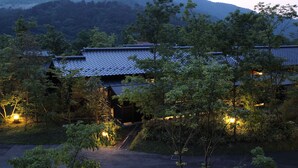 En traditionell japansk byggnad med ett tak täckt av tegelpannor, omgiven av träd och med ett bergigt landskap i bakgrunden.