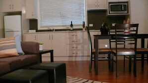 A compact living space with a sofa, ottoman, dining table, chairs, kitchen area including cabinets and microwave, and a window with blinds. - Spacious Cottage, overlooking bushland (Pokolbin)
