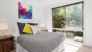 Un dormitorio con una cama grande, dos mesitas de noche, un ventanal con vistas a árboles y un cuadro abstracto de colores en la pared.