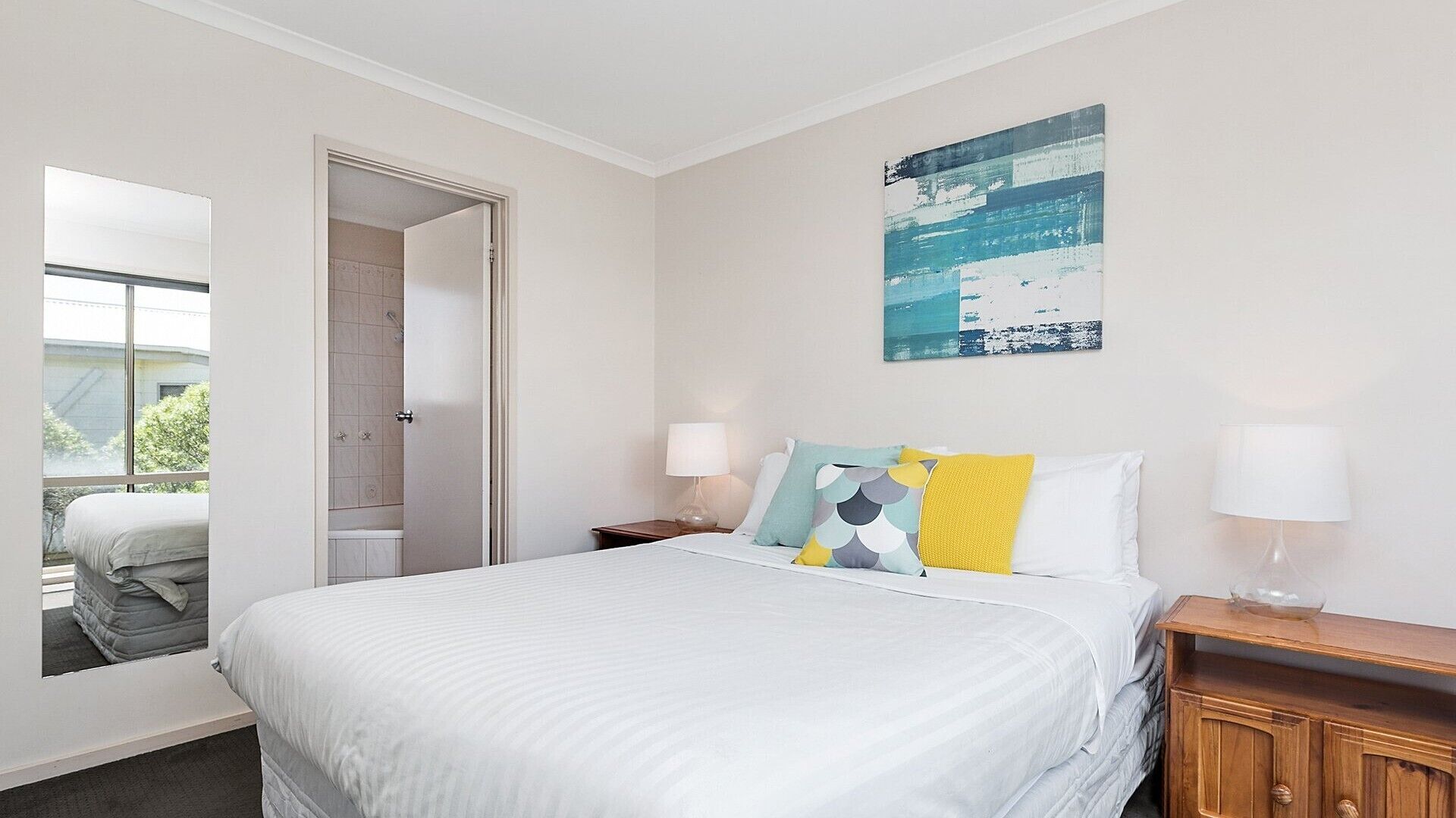 A hotel room with a bed, a nightstand, a lamp, a painting, and a view of the outside.