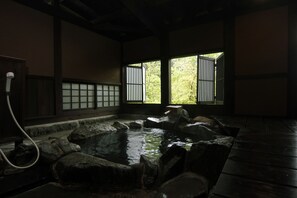 A traditional Japanese onsen with a wooden interior, a stone hot spring, and a view of greenery through a window. - Shugetsu (Kokonoe)
