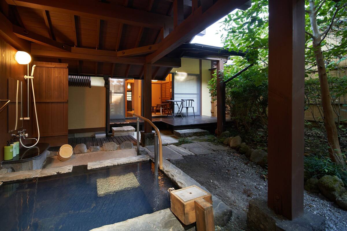 Un onsen japonés tradicional con una terraza de madera, escalones de piedra y una bañera de madera.