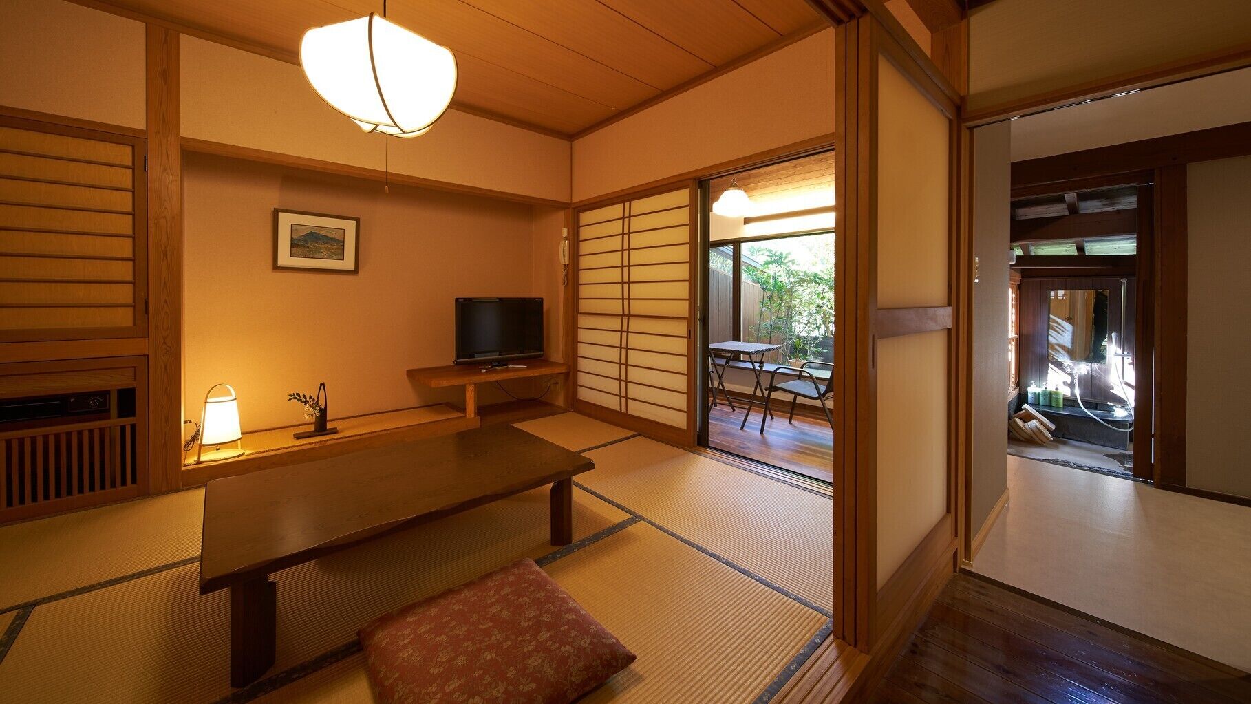 A traditional Japanese room with tatami flooring, a low table, a bench, a television, and a sliding door leading to a balcony.