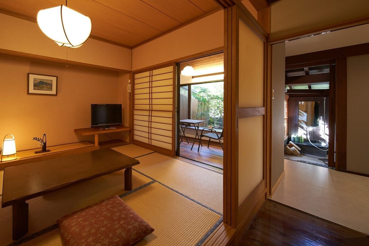 Una habitación japonesa tradicional con suelo de tatami, una mesa baja, un banco, un televisor y una puerta corrediza que da a un balcón.