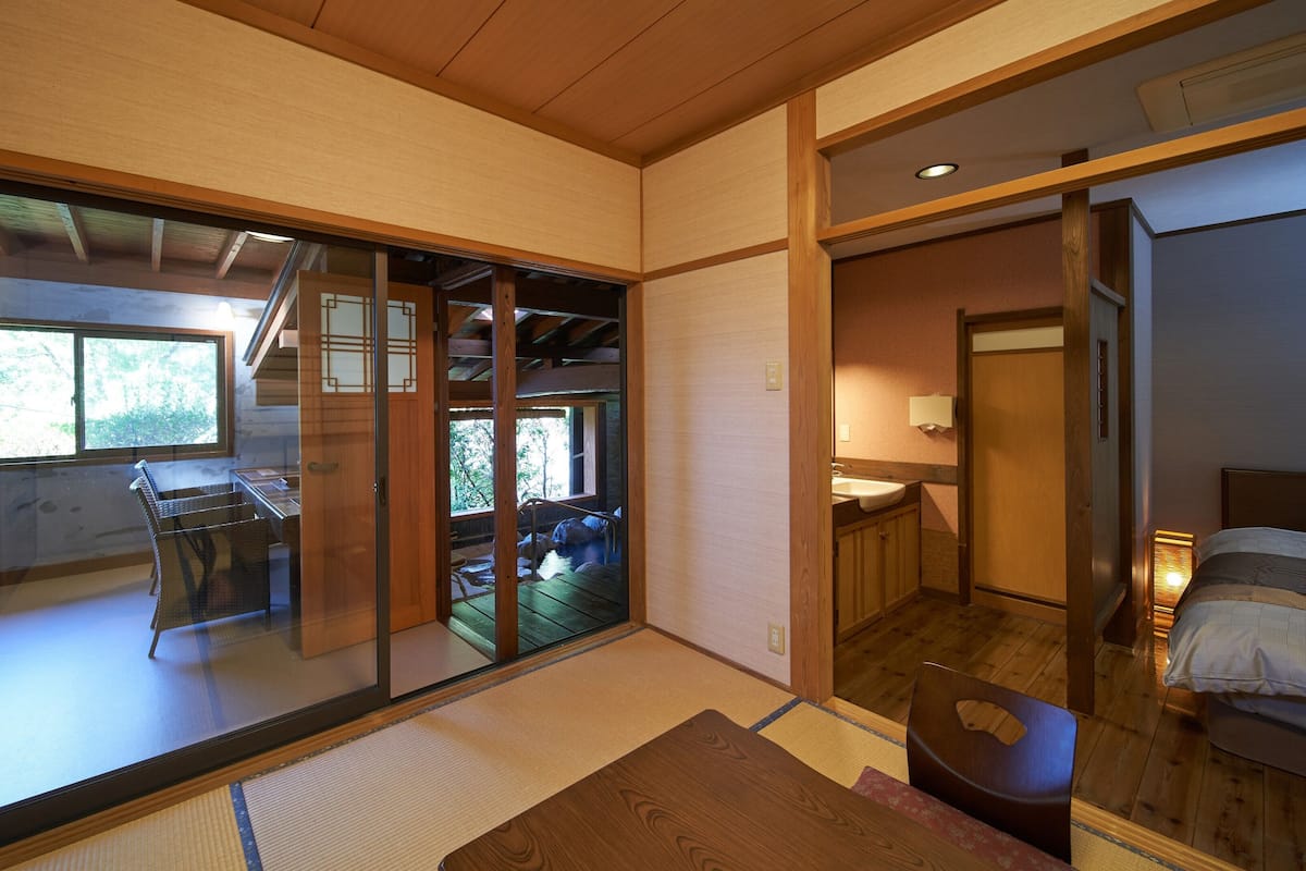 Una habitación tradicional japonesa con suelo de tatami, puertas correderas que dan a un balcón, una cama de madera y un lavabo pequeño.