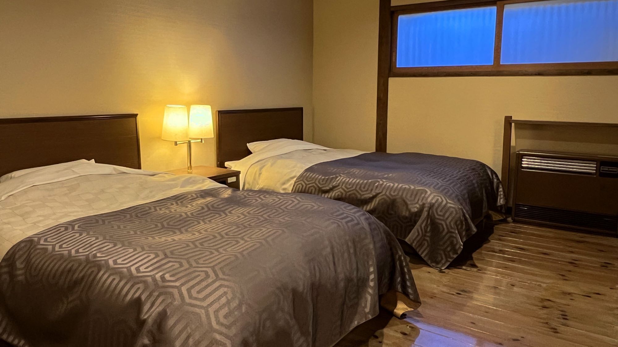 A hotel room with two beds, a nightstand with lamps, a wooden floor, and a window with blinds.