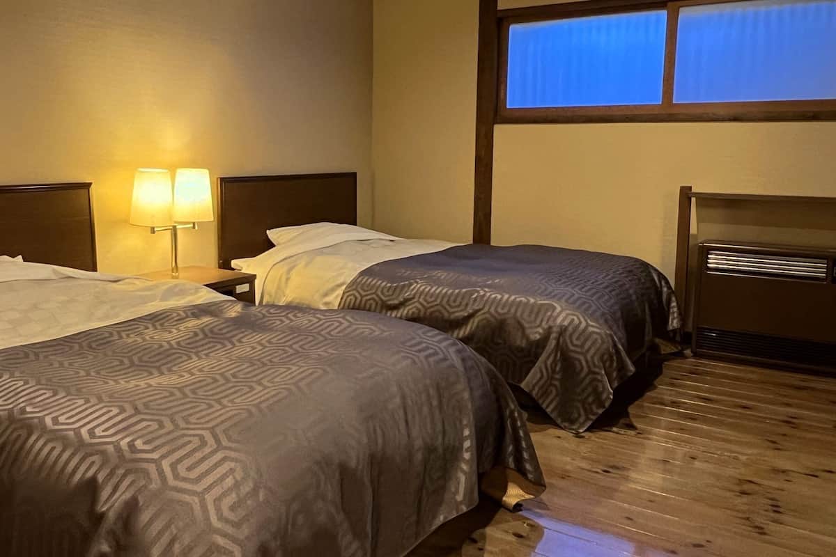 Habitación de hotel con dos camas, una mesita de noche con lámparas, piso de madera y una ventana con persianas.