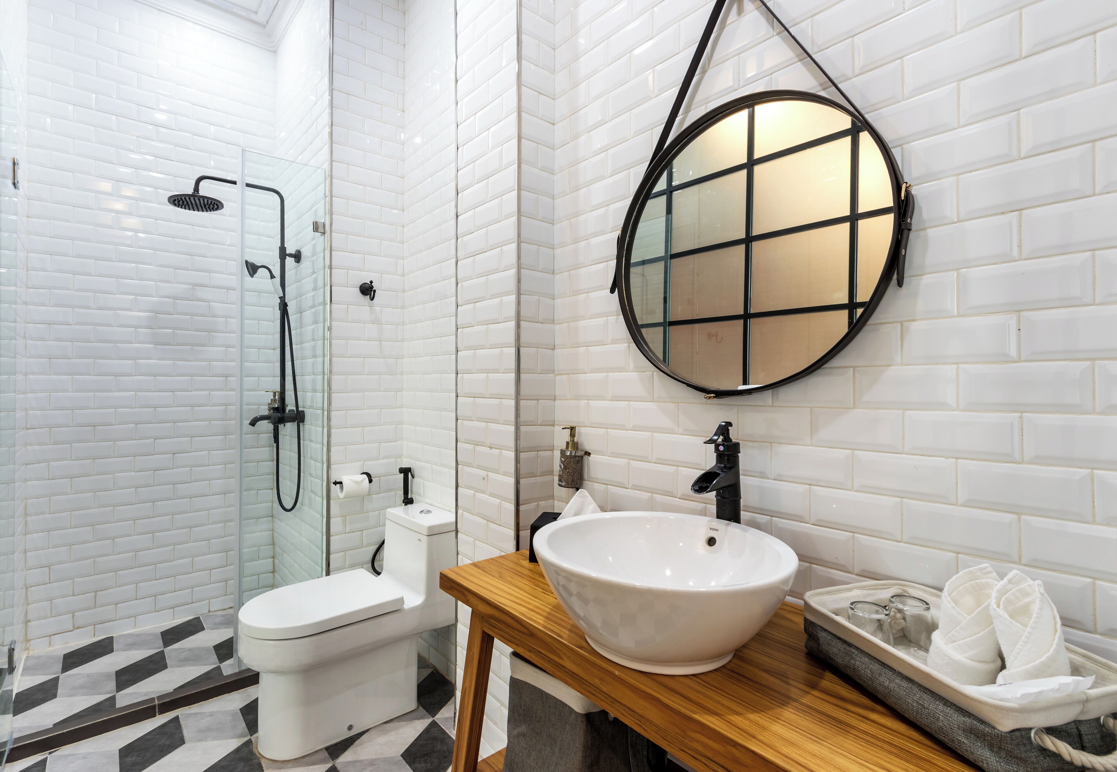 A modern bathroom with a white tiled shower, a white toilet, a wooden vanity with a white sink, and a round mirror.