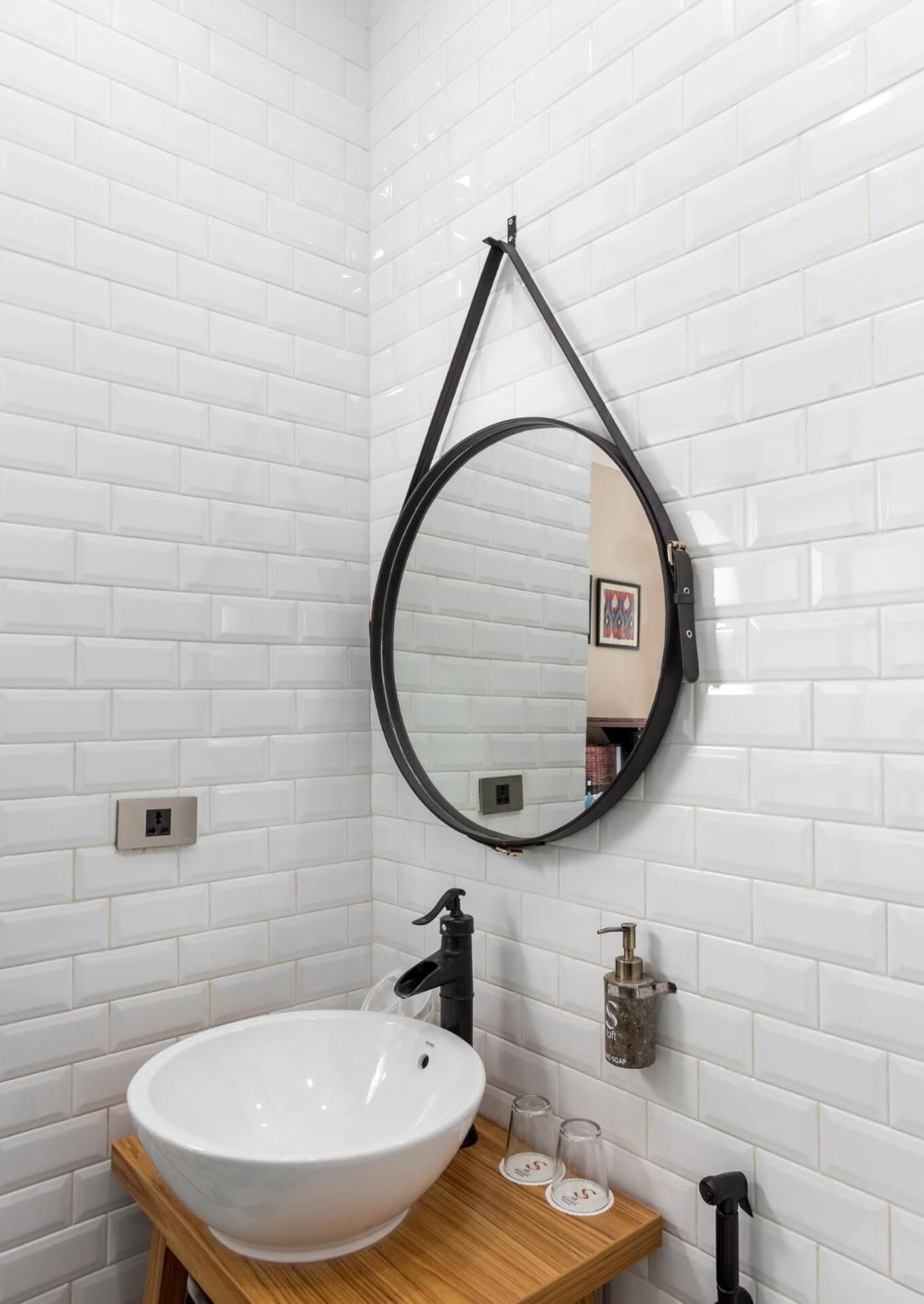 A bathroom with white tiled walls, a wooden vanity with a white sink, a black faucet, and a round mirror suspended by a black metal frame.