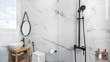 Une salle de bain moderne avec un mur en marbre blanc, un meuble-lavabo en bois, un lavabo blanc, des toilettes, une douche avec un pommeau de douche à main et un miroir.