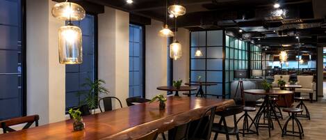 A modern dining area with a long wooden table, black chairs, hanging pendant lights, and potted plants.