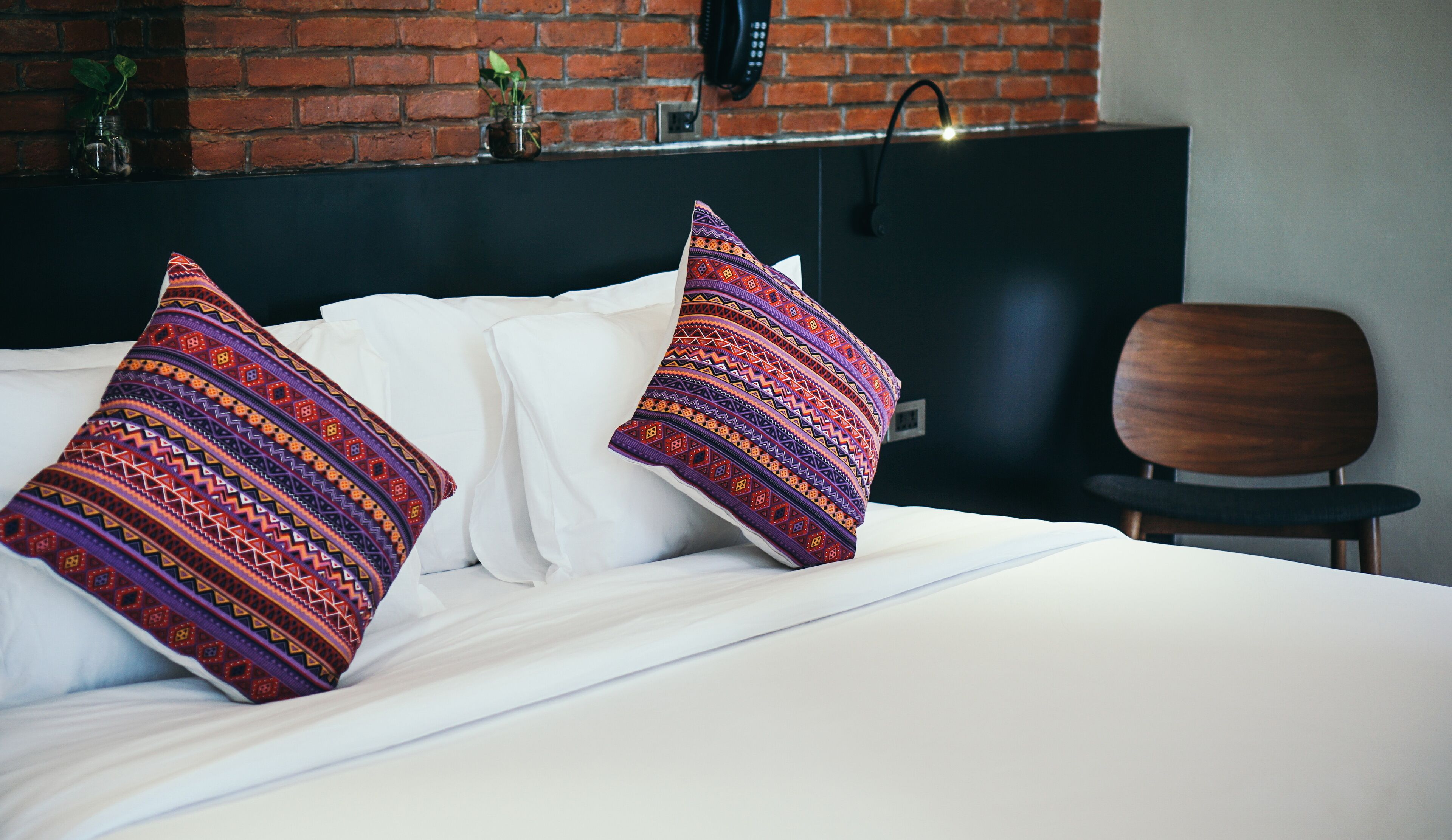 A bed with two decorative pillows, a wooden chair, and a brick wall with a phone.