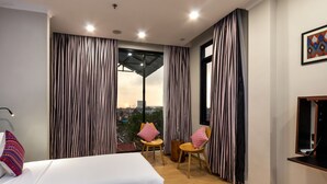 Ein modernes Hotelzimmer mit einem großen Bett, zwei Sesseln, einem kleinen Tisch und Blick auf die Stadt durch raumhohe Fenster.