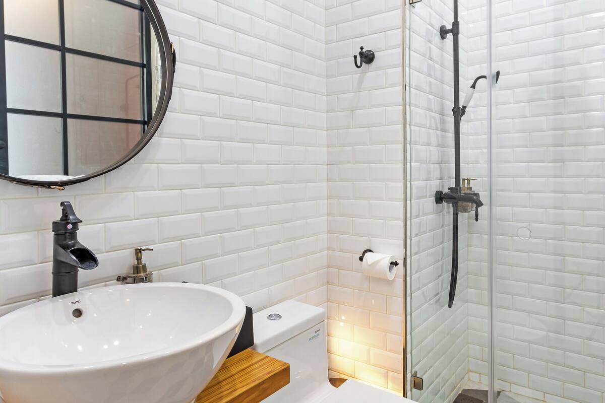 Un baño moderno con azulejos blancos tipo metro, un espejo redondo, un lavamanos blanco, un inodoro y una zona de ducha con mampara de vidrio.
