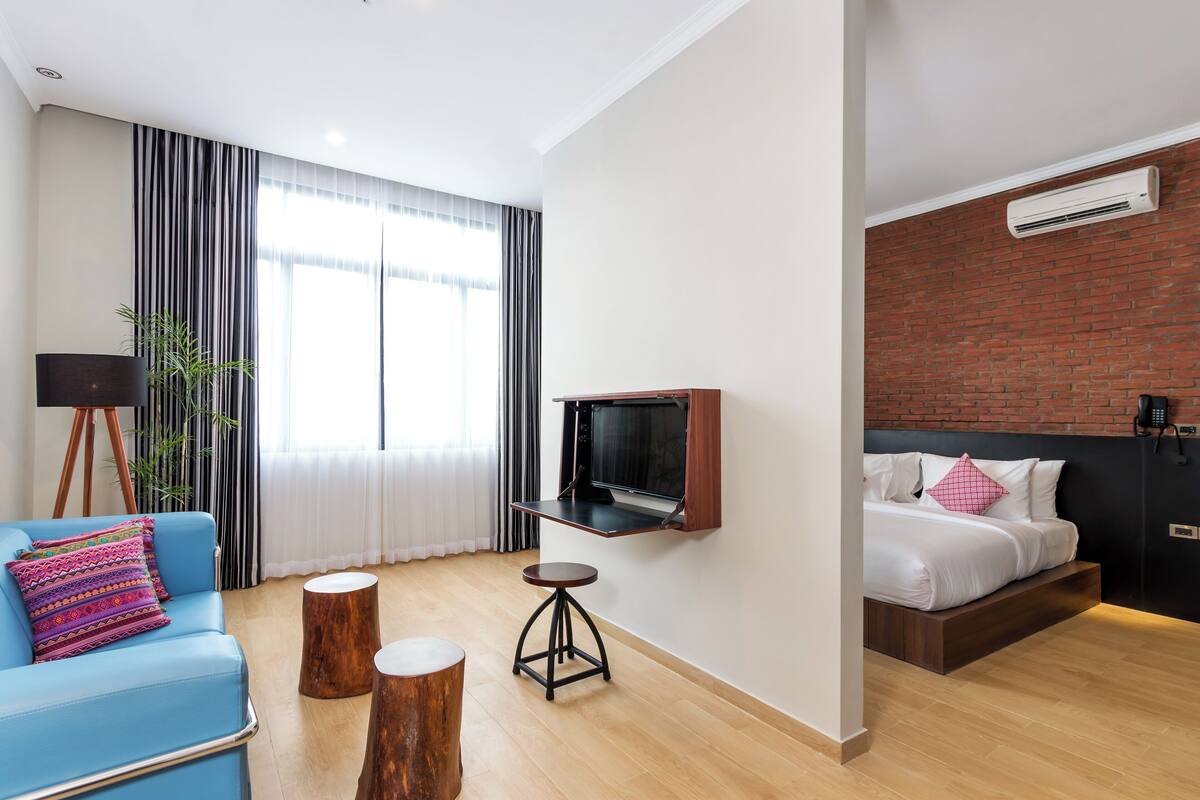 Habitación de hotel moderna con un sofá azul, un taburete de madera, un televisor montado en la pared, una cama con ropa blanca y una almohada rosa, y una pared de ladrillo decorativa.