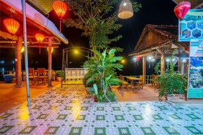 An outdoor patio area with tiled flooring, wooden tables and chairs, potted plants, and hanging lanterns. - Phong Nha Coco House (Bo Trach)