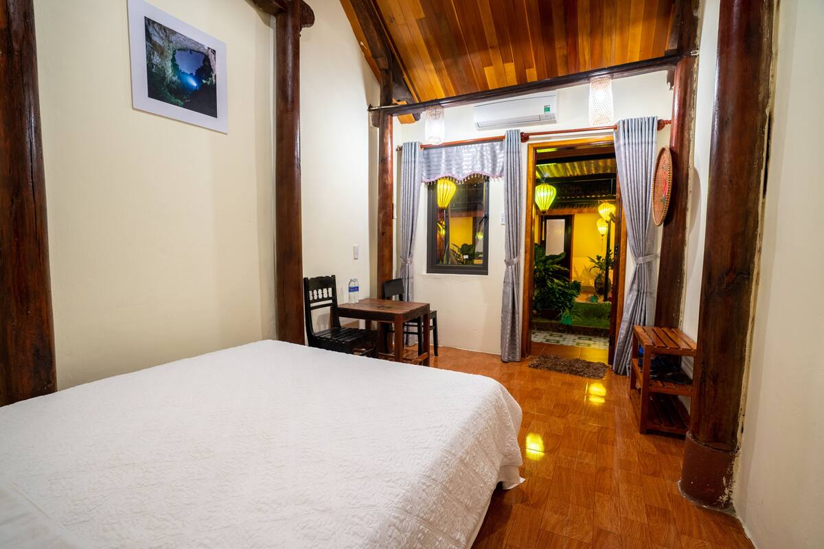 A hotel room with a bed, a wooden table, chairs, and a small desk. There is a door leading to a balcony with plants and a view.