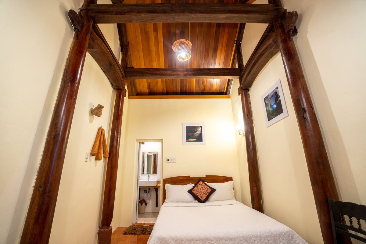 A bedroom with a wooden ceiling, a bed with white bedding, a small bathroom, and a picture on the wall.