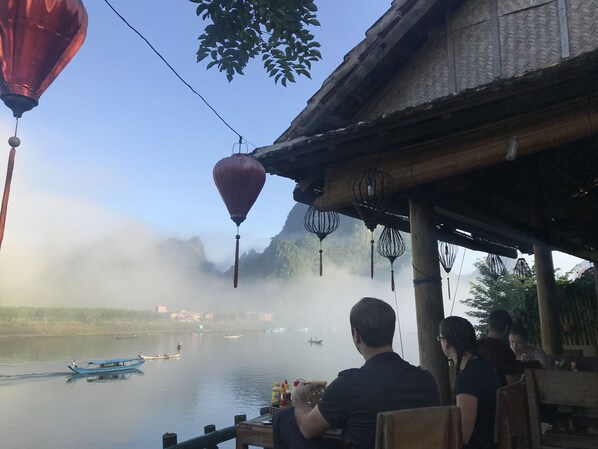 A riverside restaurant with outdoor seating, overlooking a calm river with boats. - Phong Nha Coco House (Bo Trach)