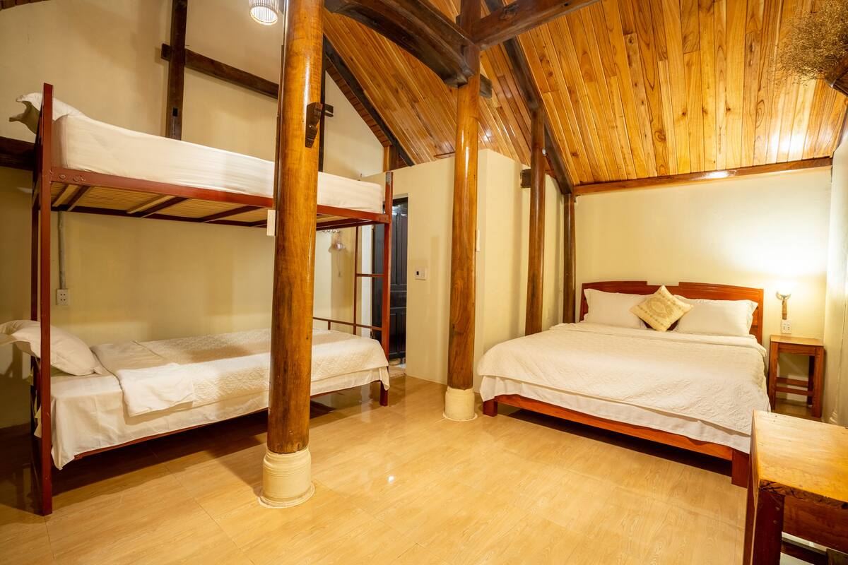 A room with two bunk beds and a single bed, wooden beams on the ceiling, and a small table with a lamp.