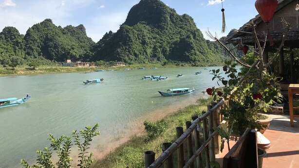 A river with boats, a mountainous backdrop, and a wooden structure with a thatched roof. - Phong Nha Coco House (Phong Nha)