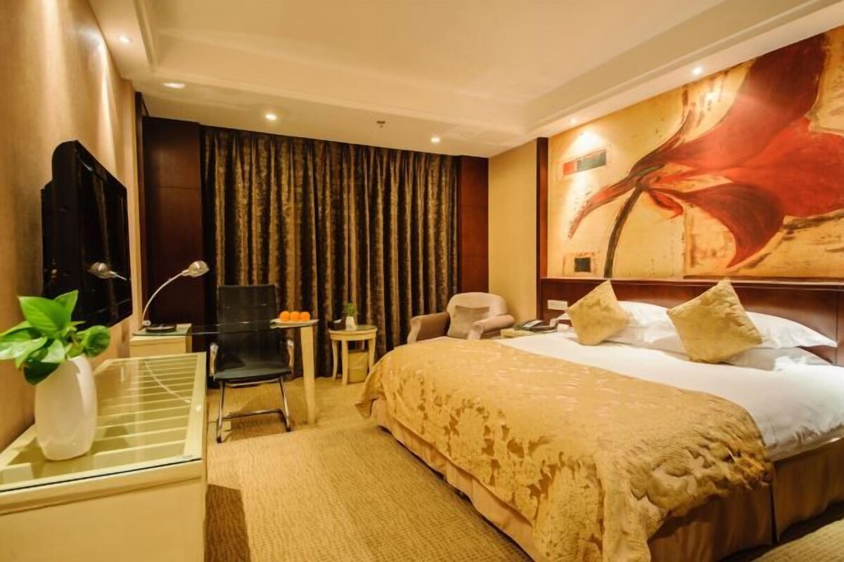 A hotel room with a large bed, a desk with a chair, a small table with a plant, and a large abstract painting on the wall.