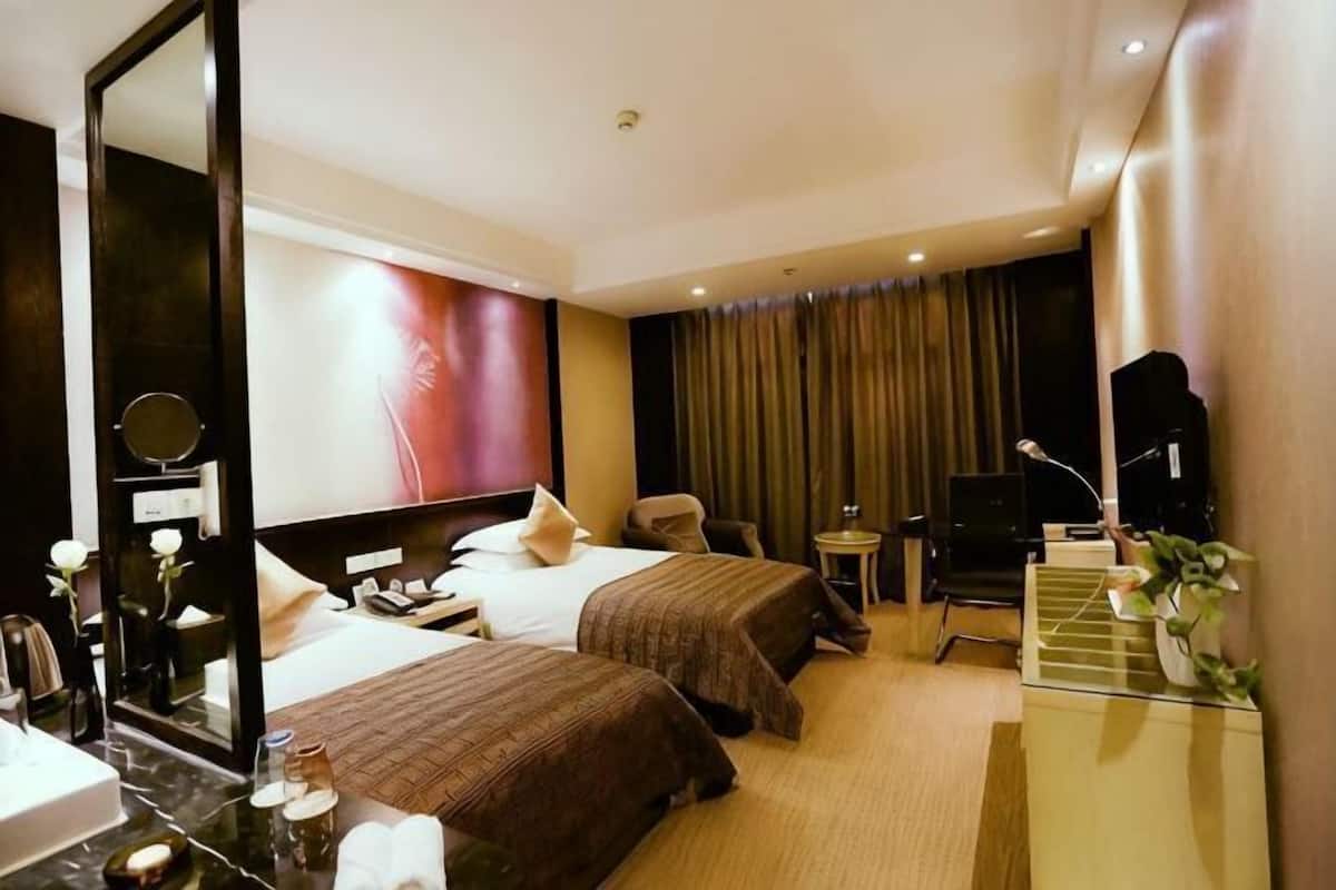A hotel room with two beds, a desk, a chair, a mirror, a TV, and a small table with a plant.