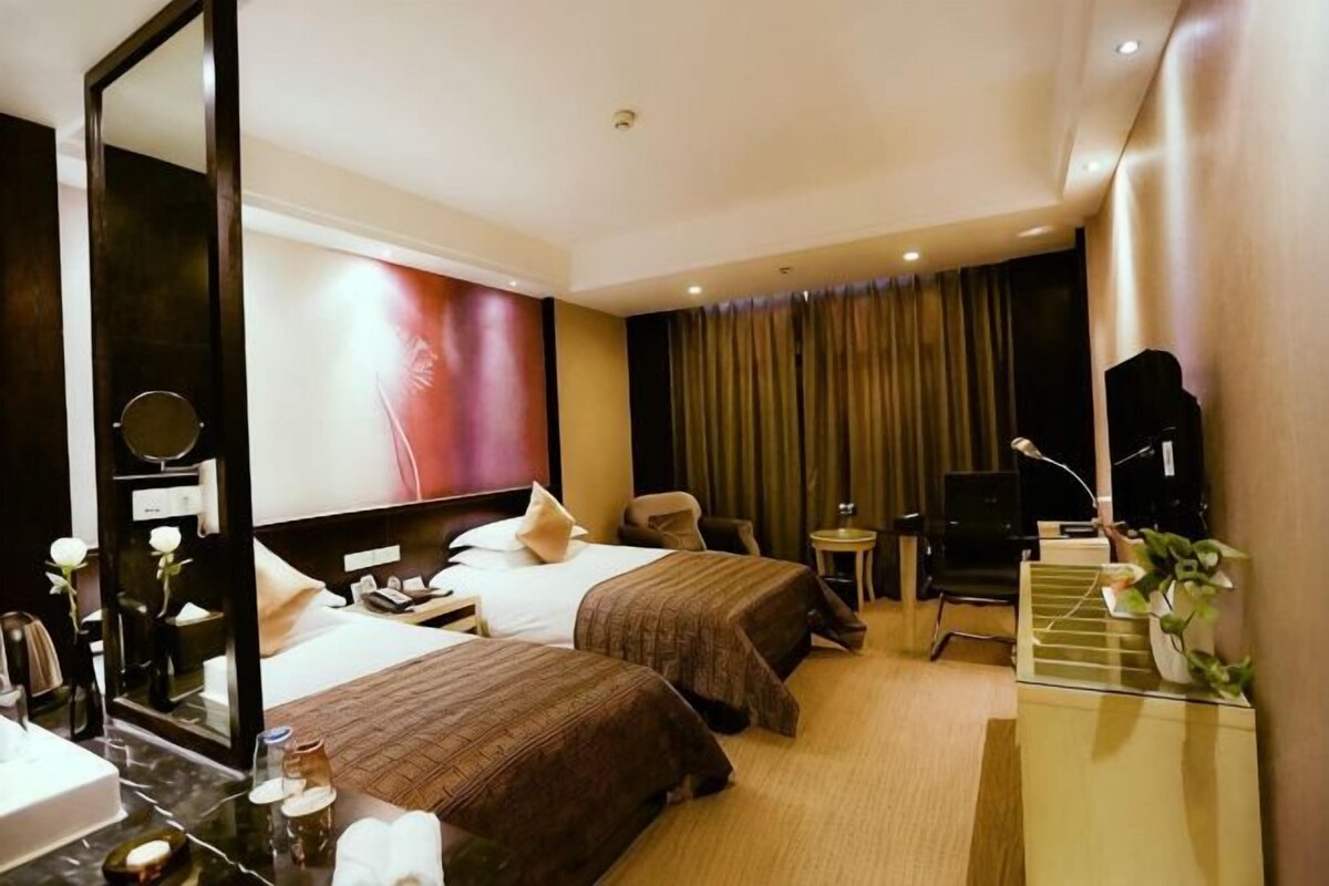 A hotel room with two beds, a desk, a chair, a mirror, a TV, and a small table with a plant.