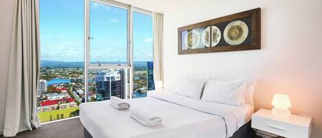 A hotel room with a large bed, a nightstand with a lamp, and a view of a cityscape through a large window.