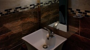 A bathroom with a white sink, a mirror, and two wall-mounted lights. The walls are tiled with a mix of brown and beige tiles. - Modern New Kingston Apartment (Secured/Parking) (Kingston)
