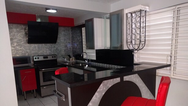 A modern kitchen with a central island, red chairs, stainless steel appliances, and a flat-screen TV mounted above the island. - Modern New Kingston Apartment (Secured/Parking) (Kingston)