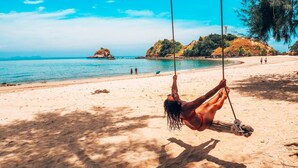 Une scène de plage avec une personne sur une balançoire, une eau d’un bleu limpide et des formations rocheuses lointaines.