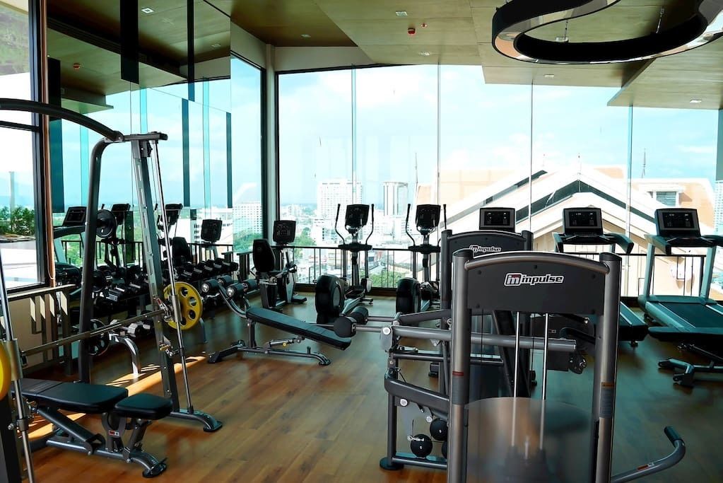 Un gimnasio con diversos equipos de ejercicio, amplias ventanas y vistas a edificios.