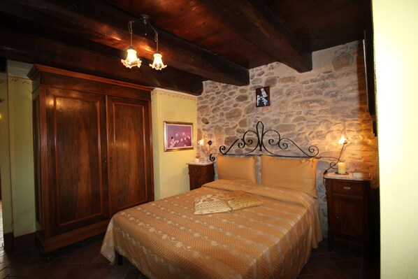 Quarto com guarda-roupa de madeira, parede de pedra, cama com roupa de cama estampada e duas mesinhas de cabeceira com velas.