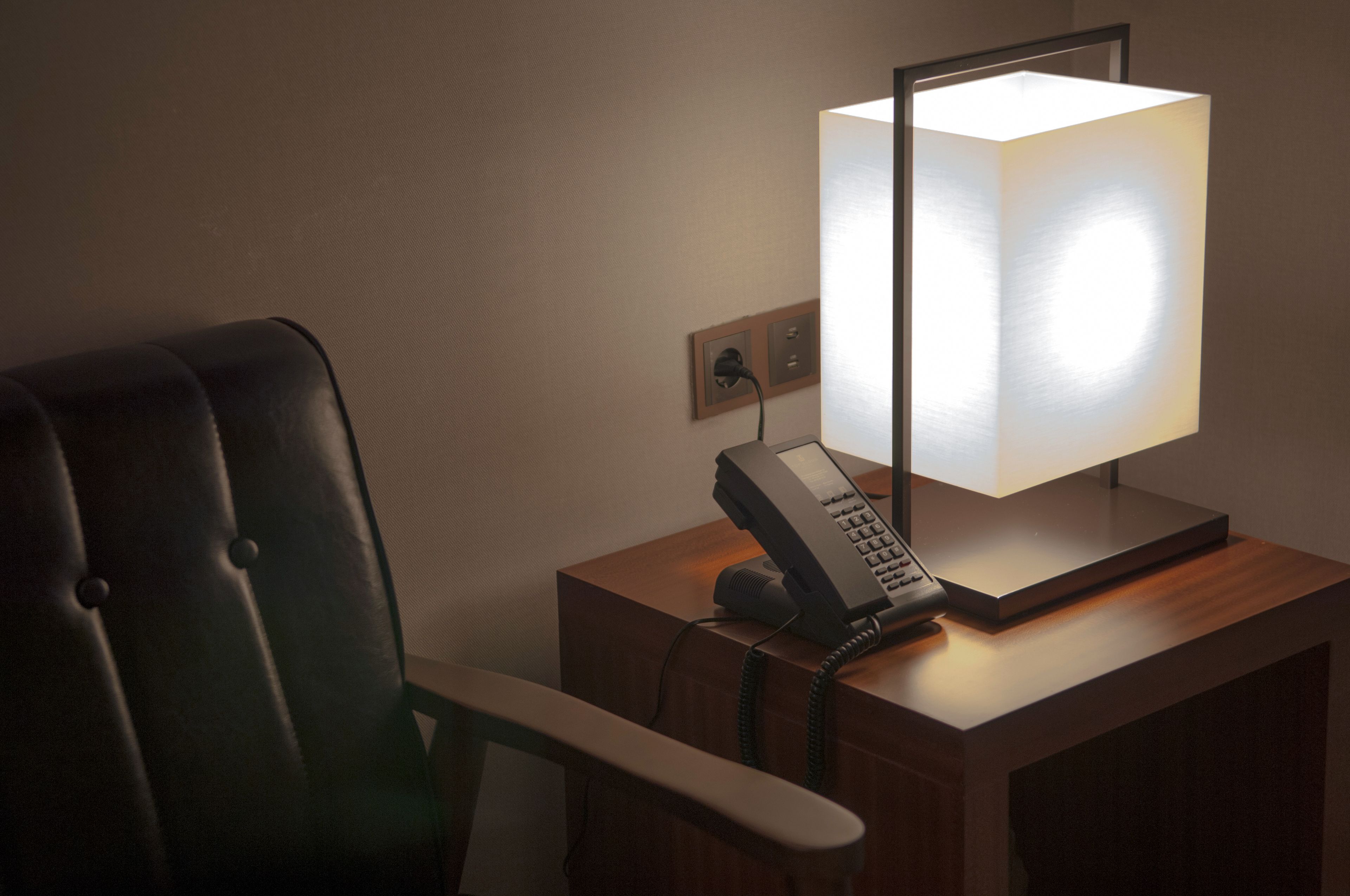 A hotel room with a wooden nightstand, a telephone, and a wall-mounted lamp.