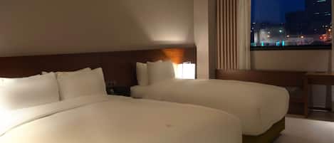 A hotel room with two large beds, a nightstand, a lamp, and a window with a city view.