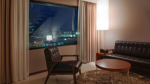 Een moderne woonkamer met een leren bank, een ronde salontafel en een fauteuil. Een groot raam biedt uitzicht op een stadsbeeld 's nachts.