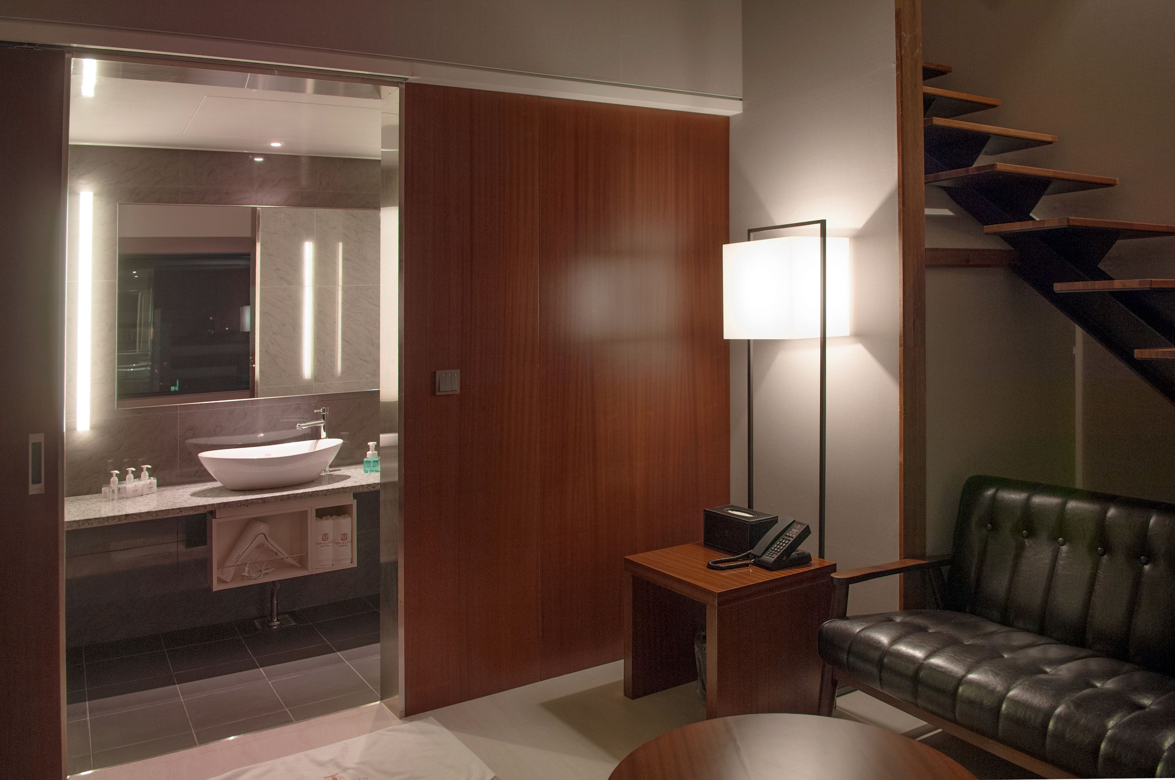 A modern bathroom with a large mirror, a sink, and a vanity. There is a wooden door, a floor lamp, and a leather sofa in the adjacent living area.