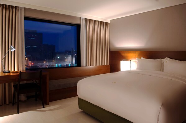 A hotel room with a large bed, a desk with a chair, and a view of a cityscape at night.