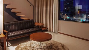 Een moderne woonkamer met een leren bank, een ronde salontafel, een klein houten tafeltje en uitzicht op een stadsbeeld door een groot raam.