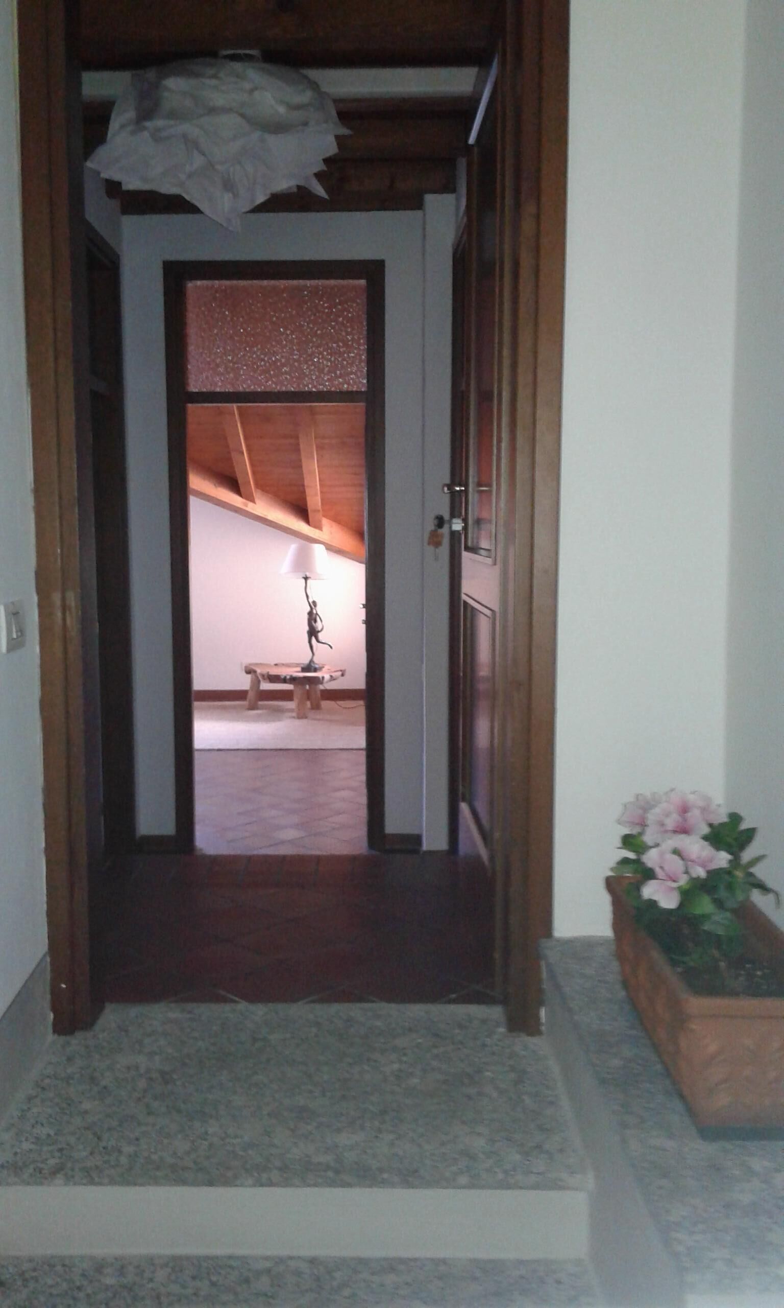 Un couloir avec une porte en bois qui mène à une pièce contenant un lit et une table. Une lampe est posée sur la table, et un pot de fleurs se trouve sur la droite.