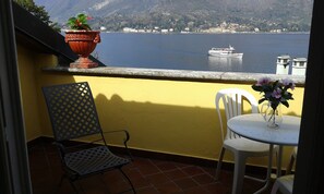 Un balcon avec un mur jaune, une plante en pot, une chaise blanche, une petite table avec un vase de fleurs et une vue sur un lac avec un bateau.
