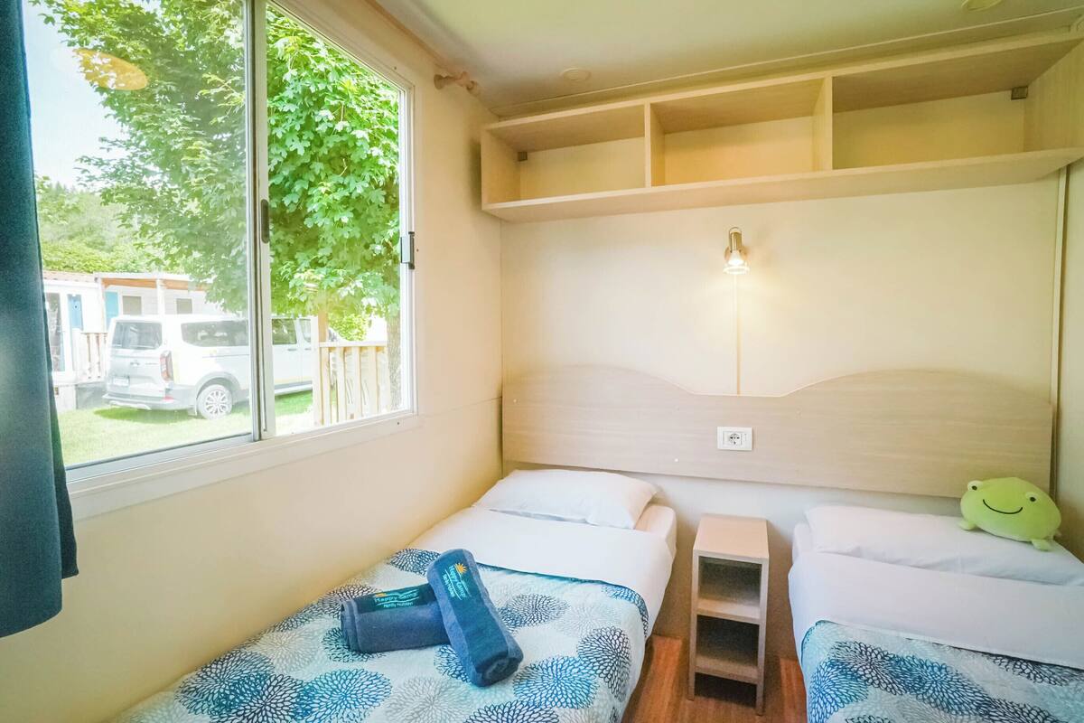 A small, cozy room with two beds, a window with a view of the outside, and a shelf above the beds.