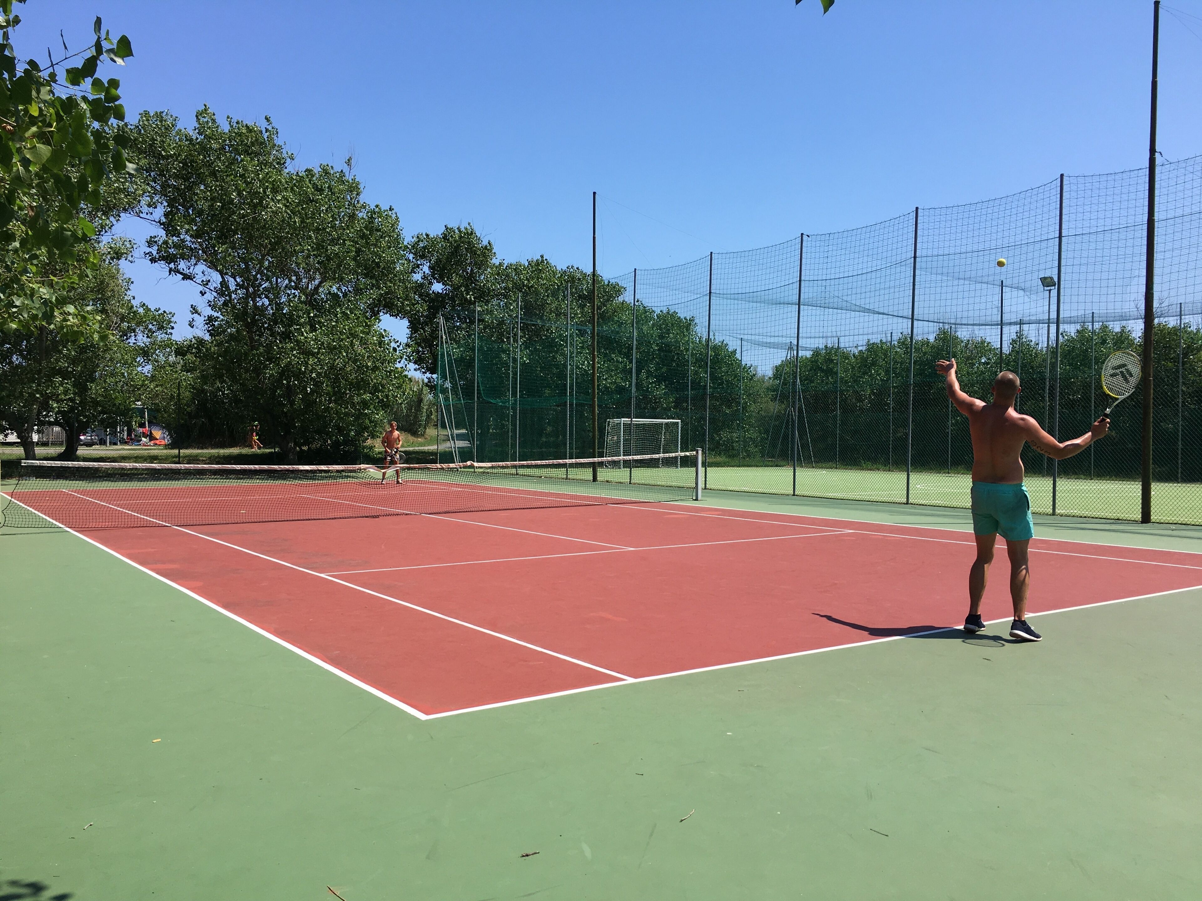 An outdoor tennis court with a net, surrounded by trees and a fence.