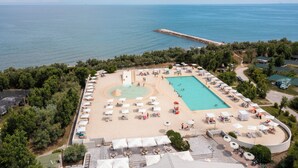 Vue aérienne d’un complexe hôtelier en bord de mer, avec une piscine, des chaises longues et une jetée qui s’avance dans la mer.