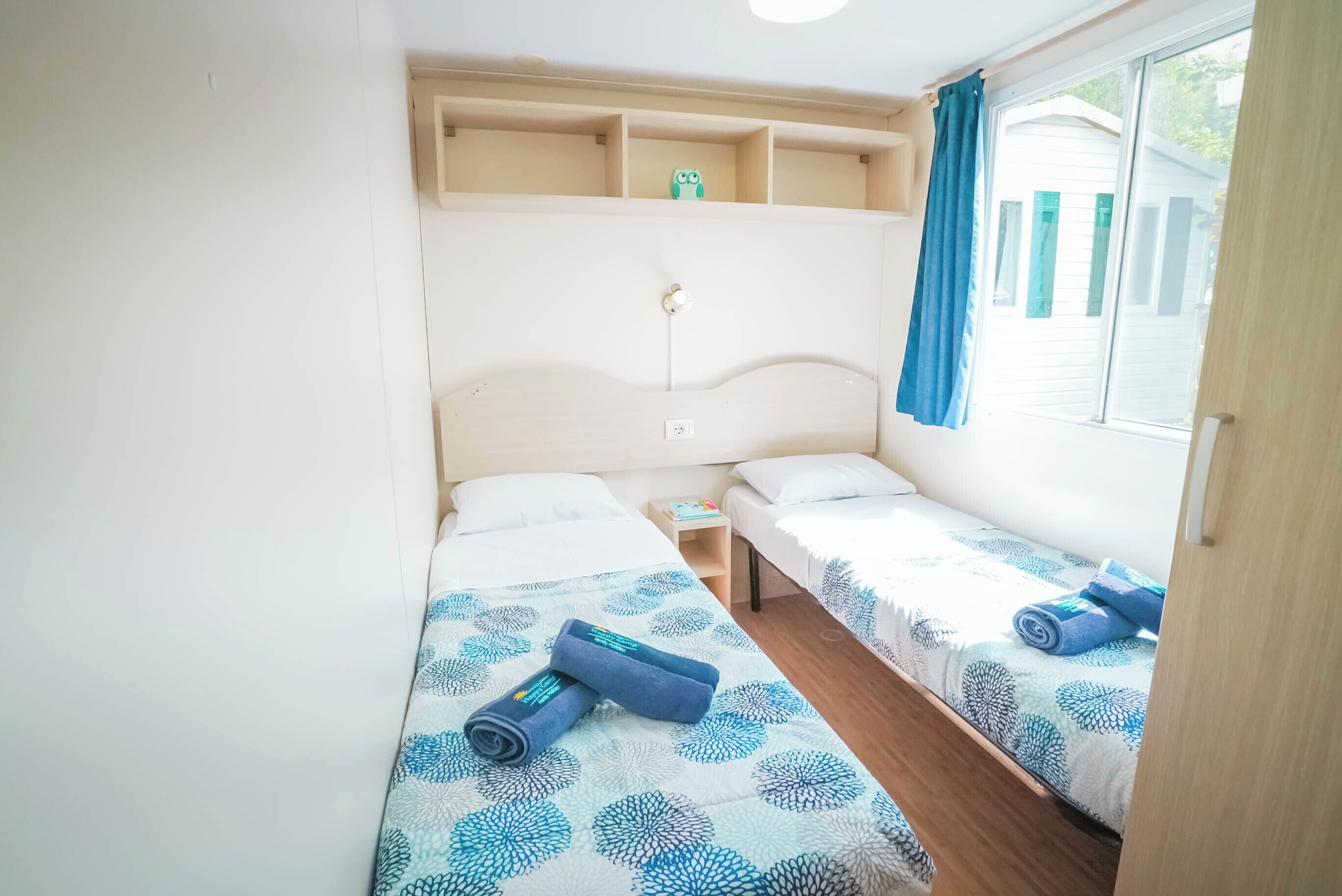 A small, well-lit room with two single beds, a wooden headboard, a shelf with a small toy, and a window with curtains.