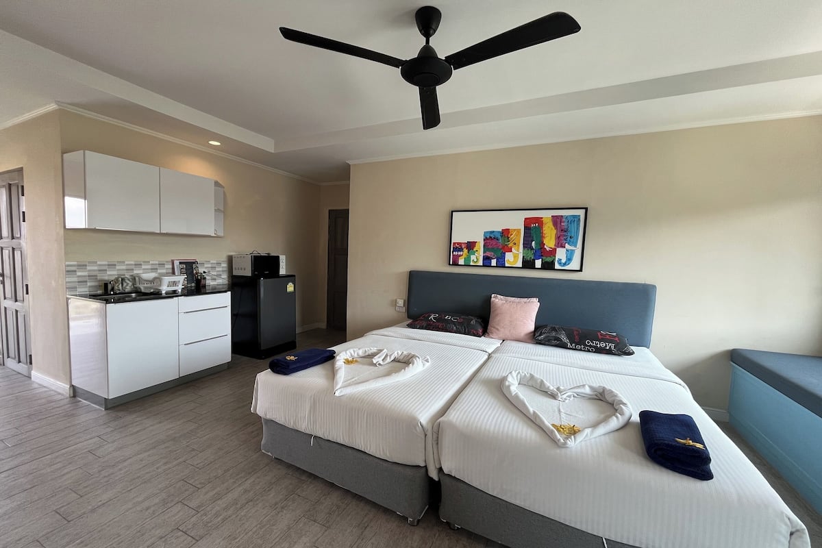 Une chambre d’hôtel avec un lit, un ventilateur de plafond, une kitchenette et une peinture abstraite aux couleurs vives accrochée au mur.